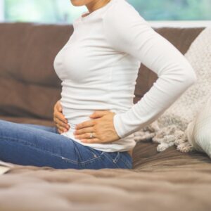 Woman holding her abdomen, expressing pain, while sitting on the couch.
